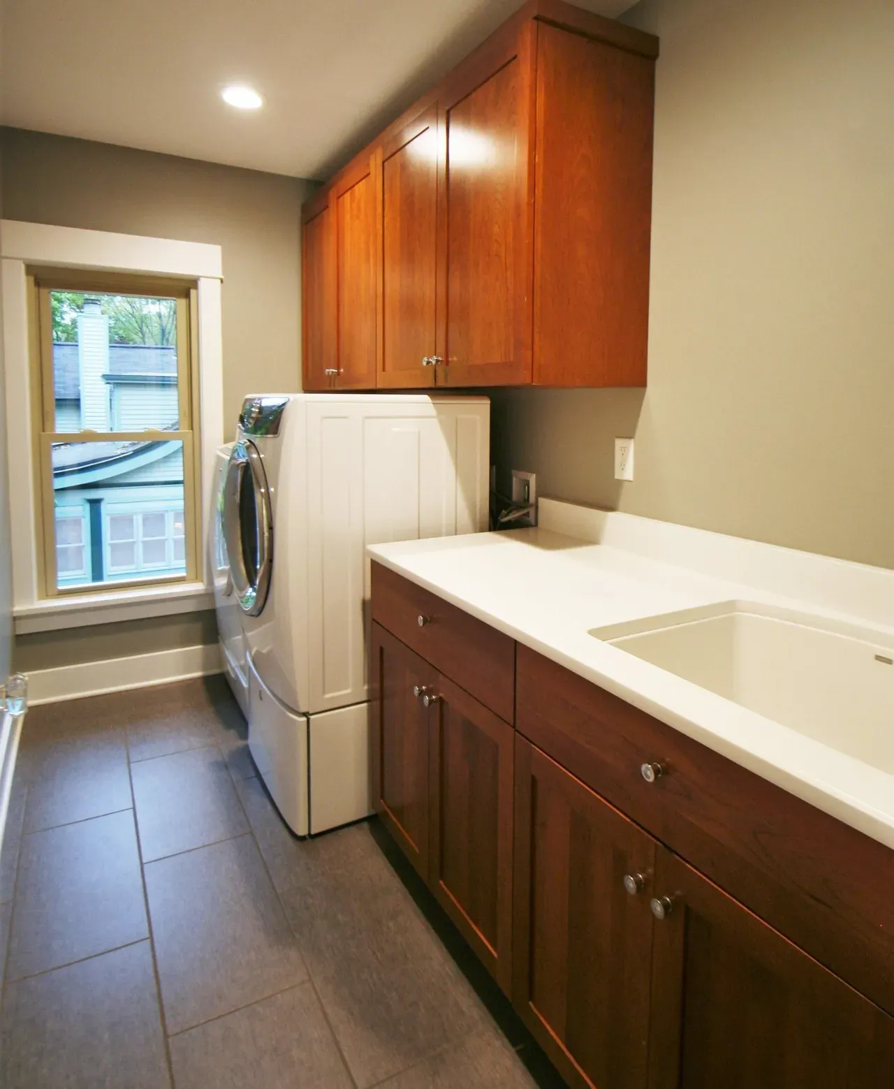 Narrow laundry room with wood cabinets, white countertop sink, stacked washer-dryer, and window