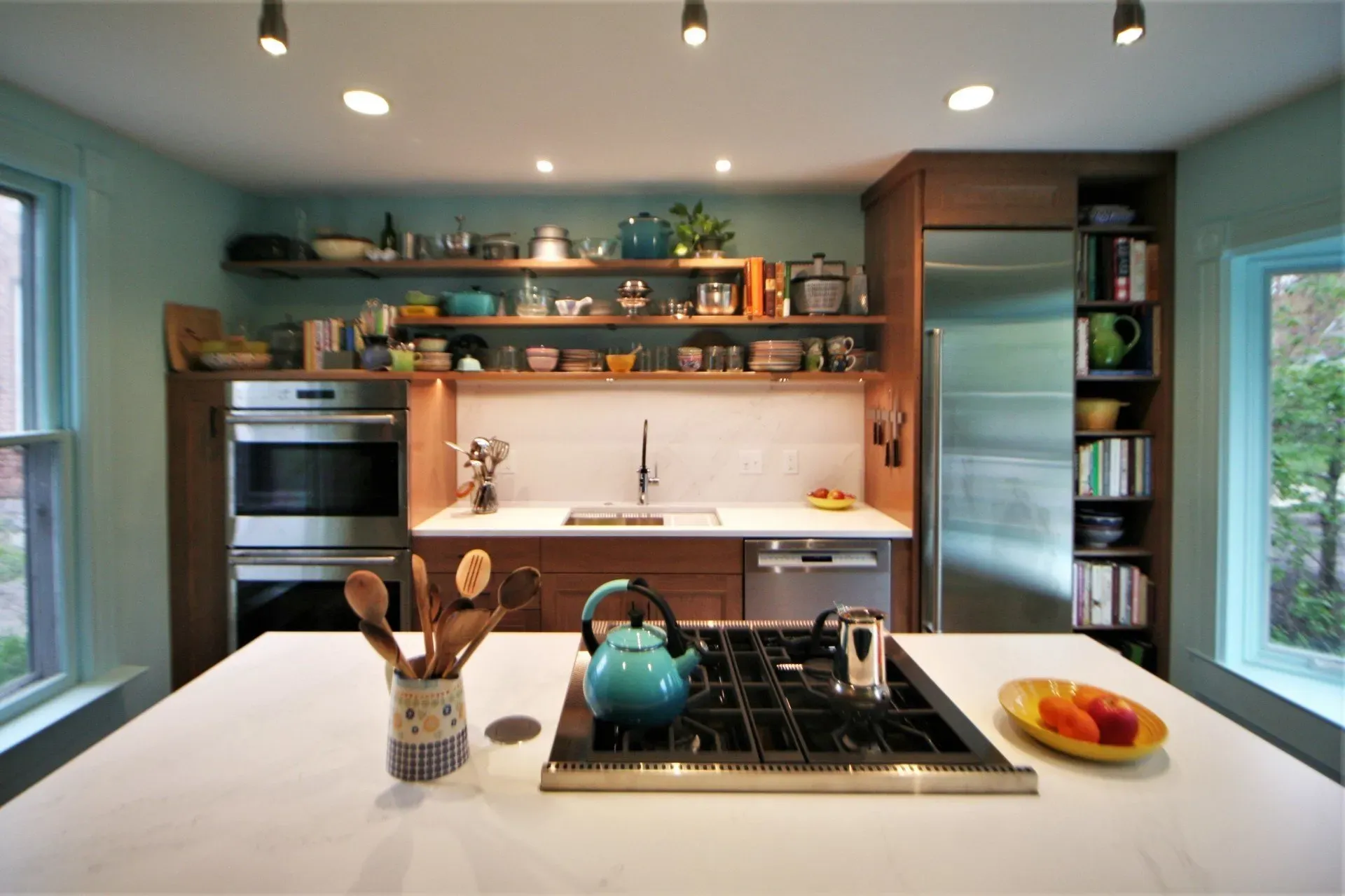 Modern kitchen interior with teal walls, wood shelves, white island, and a stovetop centerpiece.