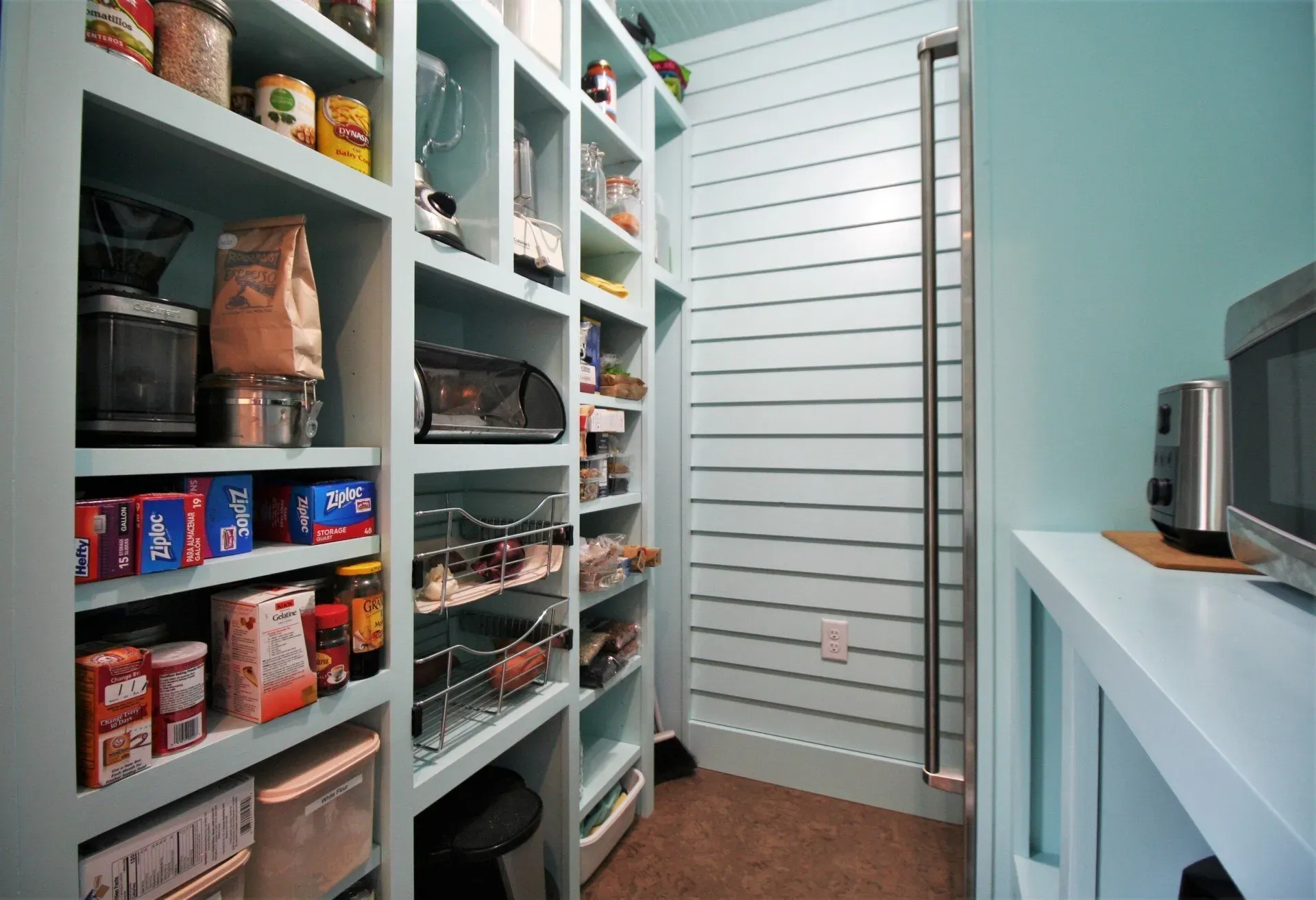Walk-in pantry with white shelves stocked with food and appliances, next to a closed door and countertop