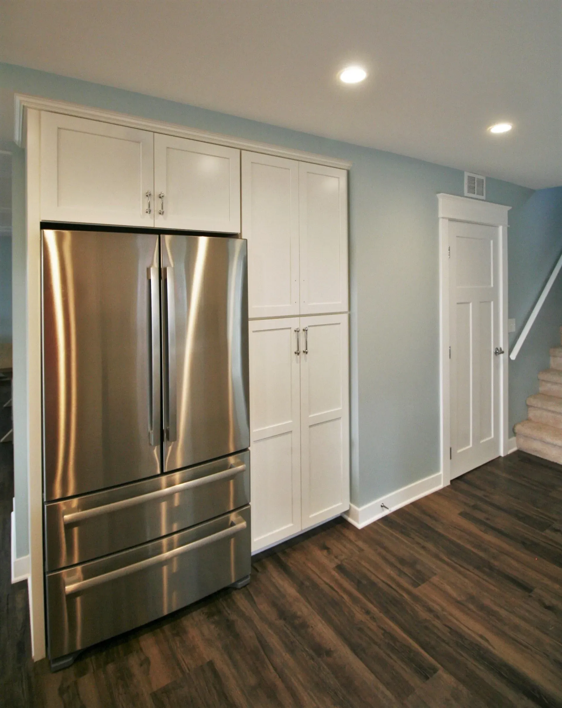 Modern kitchen with stainless steel refrigerator, white cabinets, dark wood floor, and staircase at right