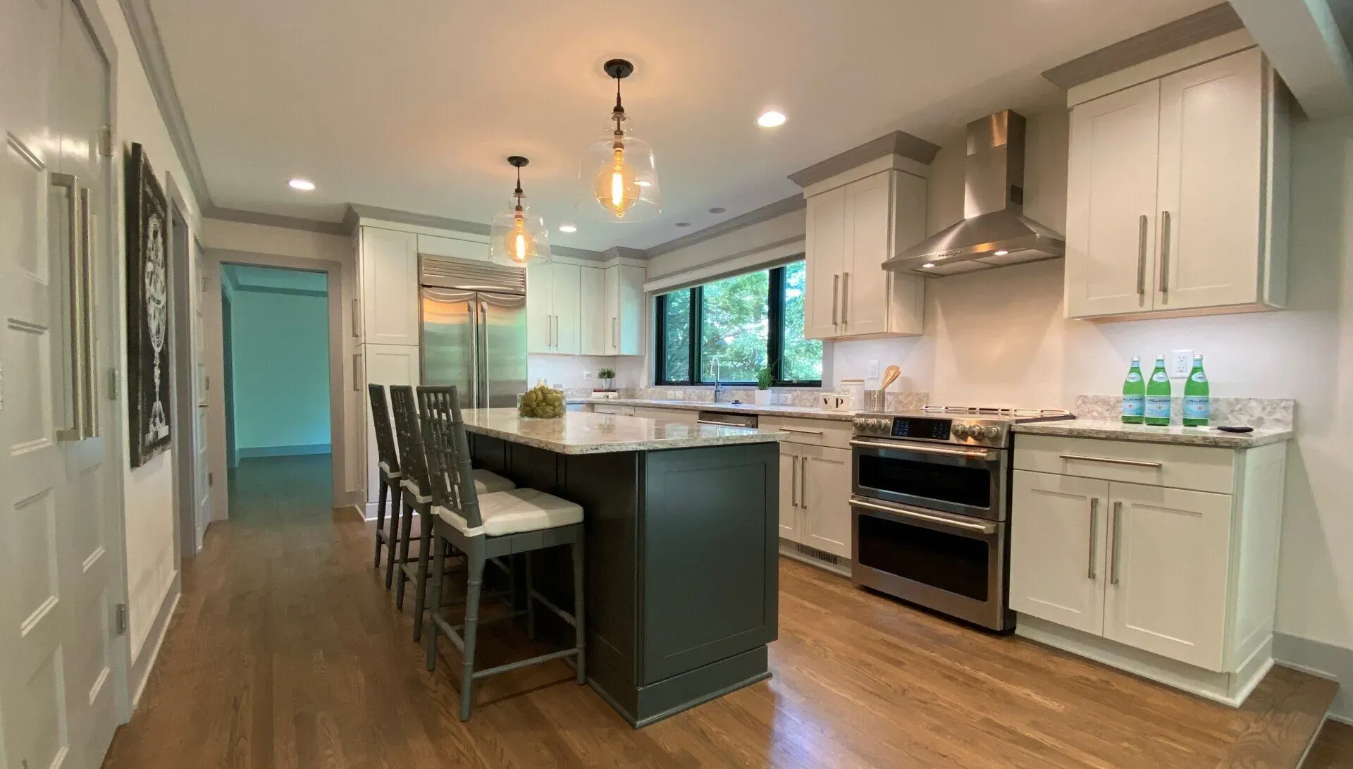 Modern kitchen with island, white cabinets, hardwood floors, and stainless steel appliances; bright windows and pendant lights