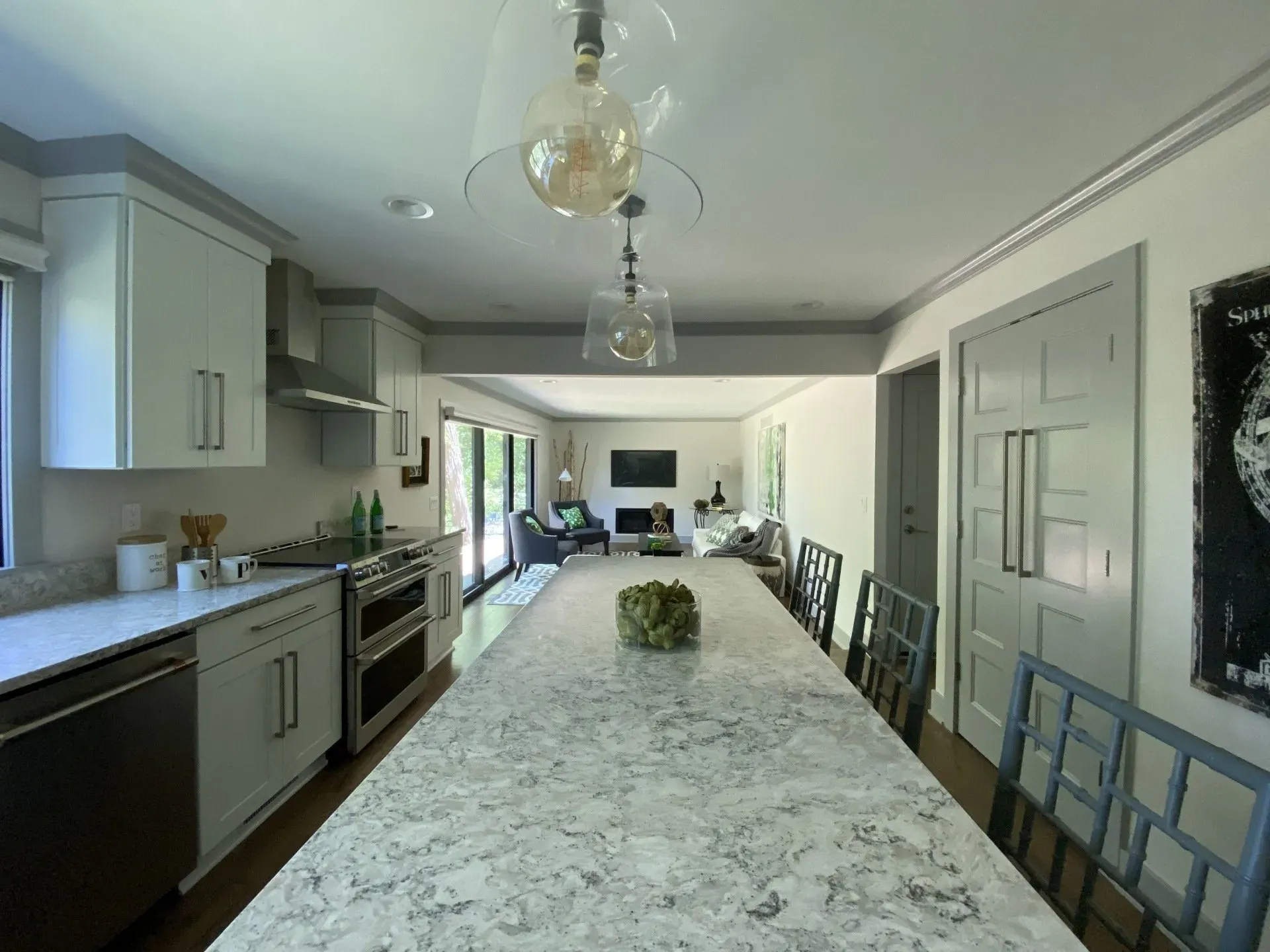 Long kitchen with a marble island, gray cabinets, stainless appliances, and chairs along the right wall.