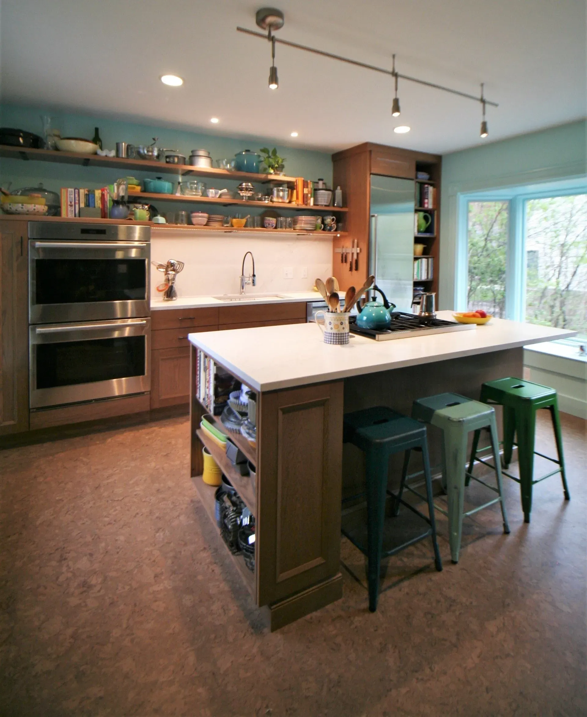 Modern kitchen with island, teal walls, wood shelves, and large window overlooking greenery