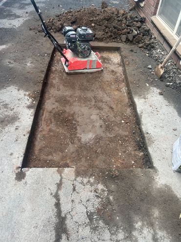 A trench being dug in concrete, with a red compact machine at the far end and dirt piled around it. A trench being dug in concrete, with a red compact machine at the far end and dirt piled around it.