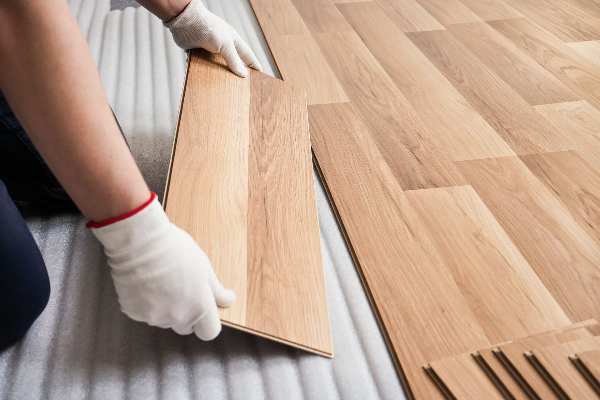 Gloved hands installing light wood laminate flooring over underlayment