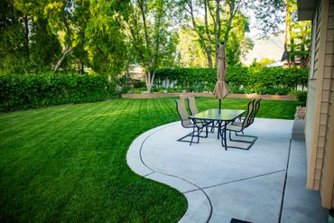 Patio with metal table and chairs beside a curved concrete path and green lawn in a sunny garden Patio with metal table and chairs beside a curved concrete path and green lawn in a sunny garden