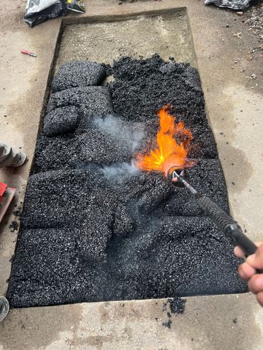 Hand torch heating a patch of black aggregate on a rectangular board outdoors.