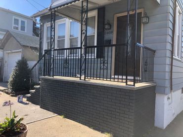 Small front porch with black railing on a light gray house, accessed by concrete steps and driveway. Small front porch with black railing on a light gray house, accessed by concrete steps and driveway.