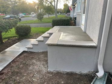 Front steps and raised concrete porch beside a house, with a small yard and sidewalk.