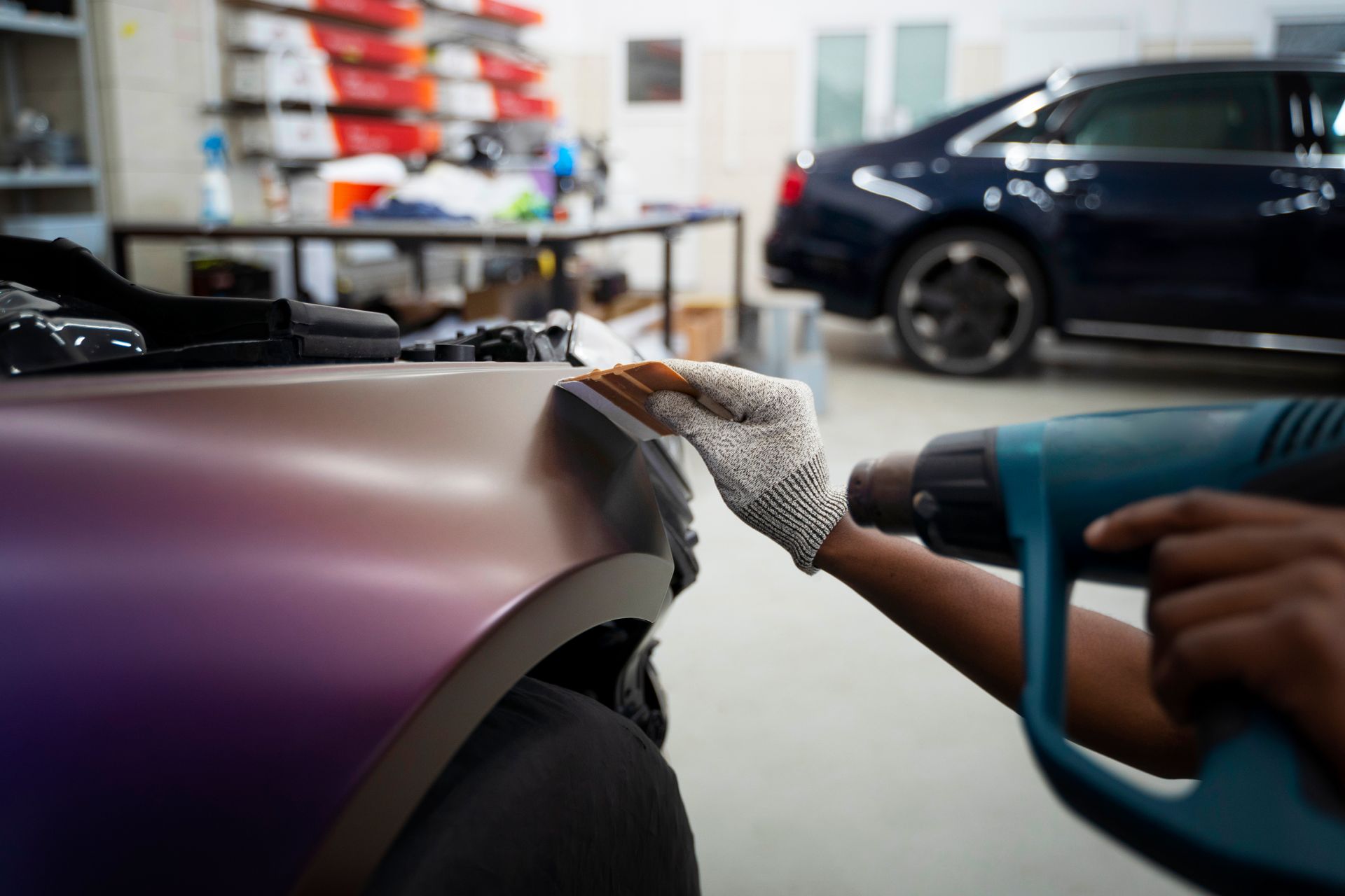 Person polishing a purple car fender in a garage with a cloth and handheld heat gun