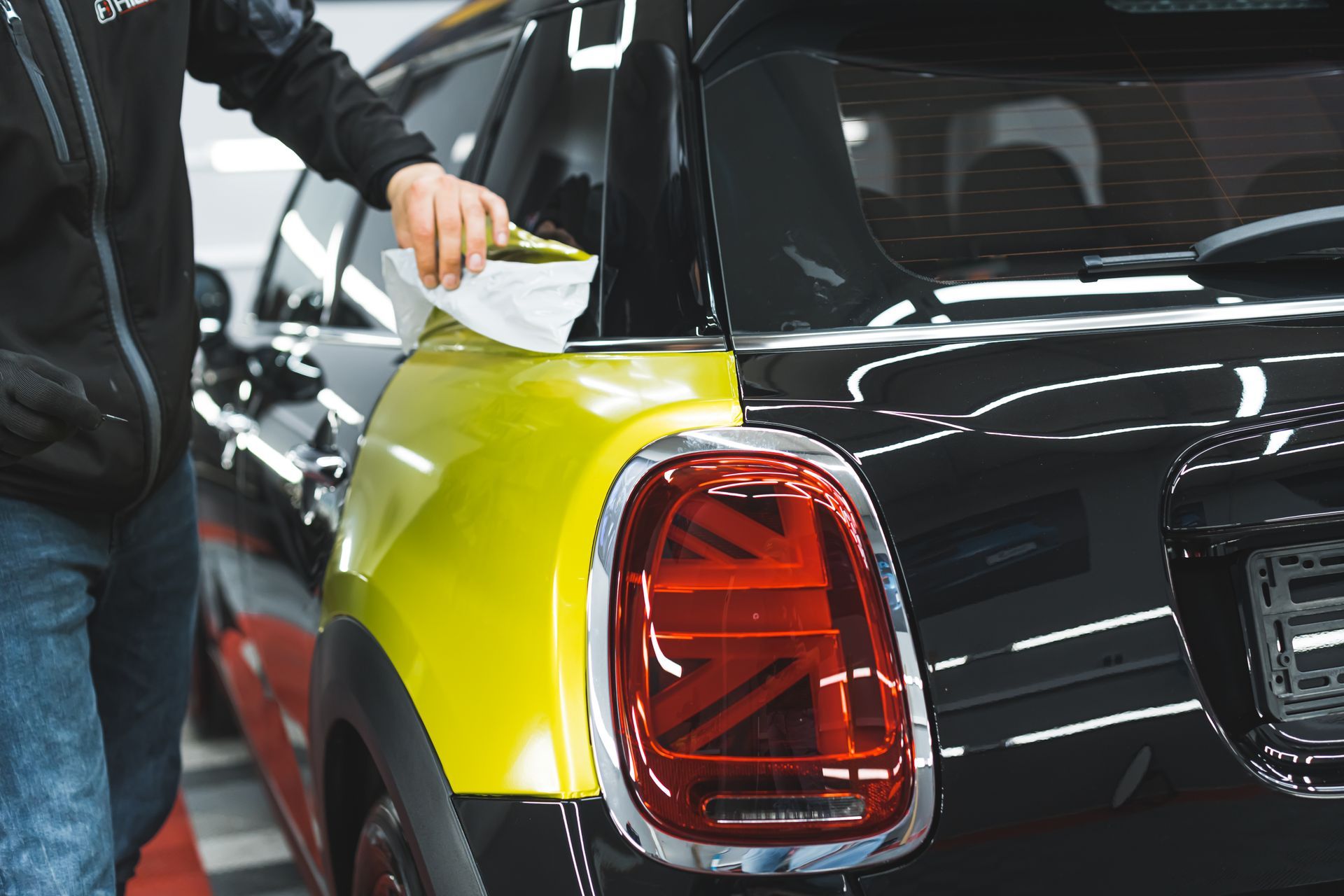 Person wiping a yellow car panel beside a red taillight in a garage
