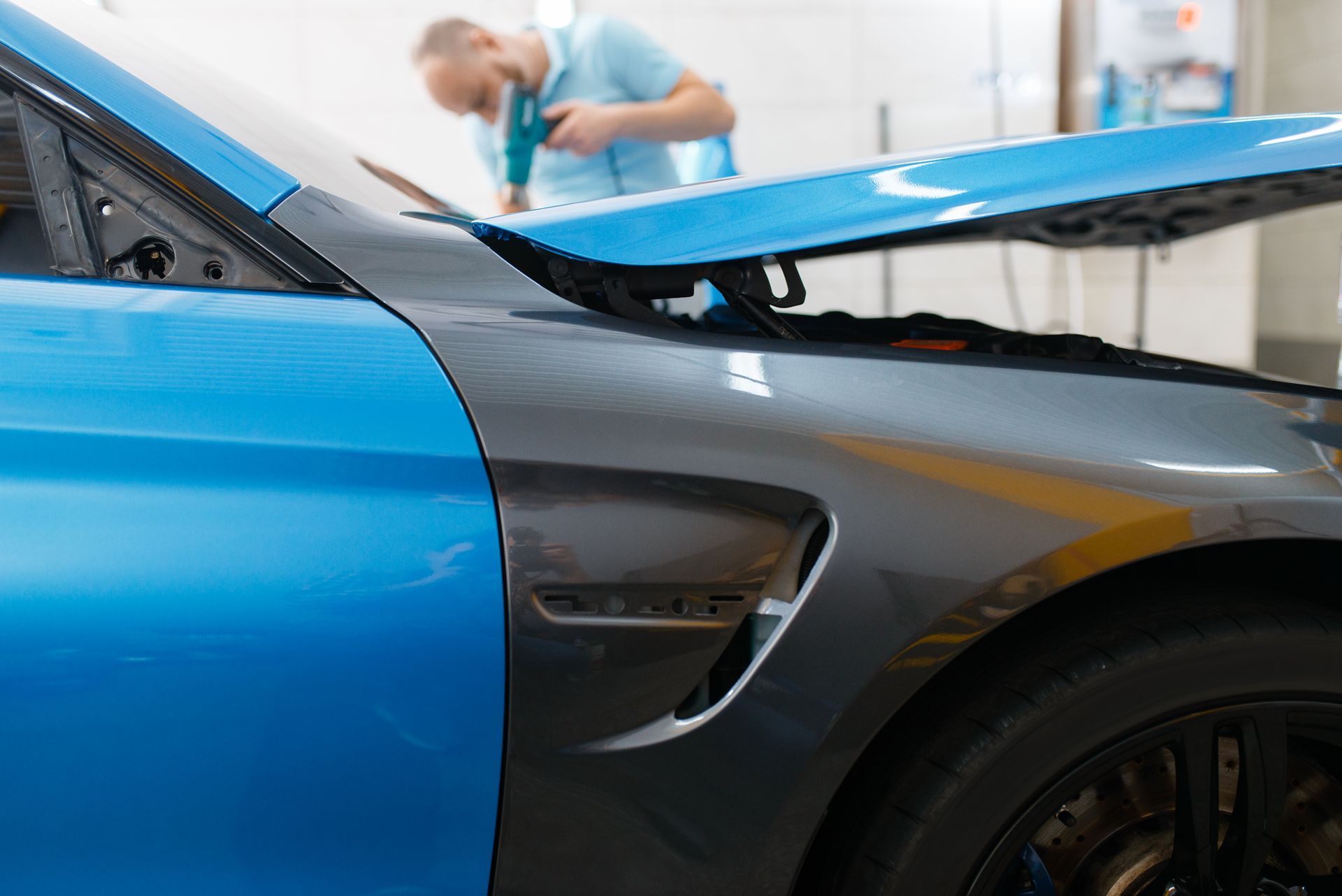 Blue sports car with raised hood in a garage, with a mechanic working in the background