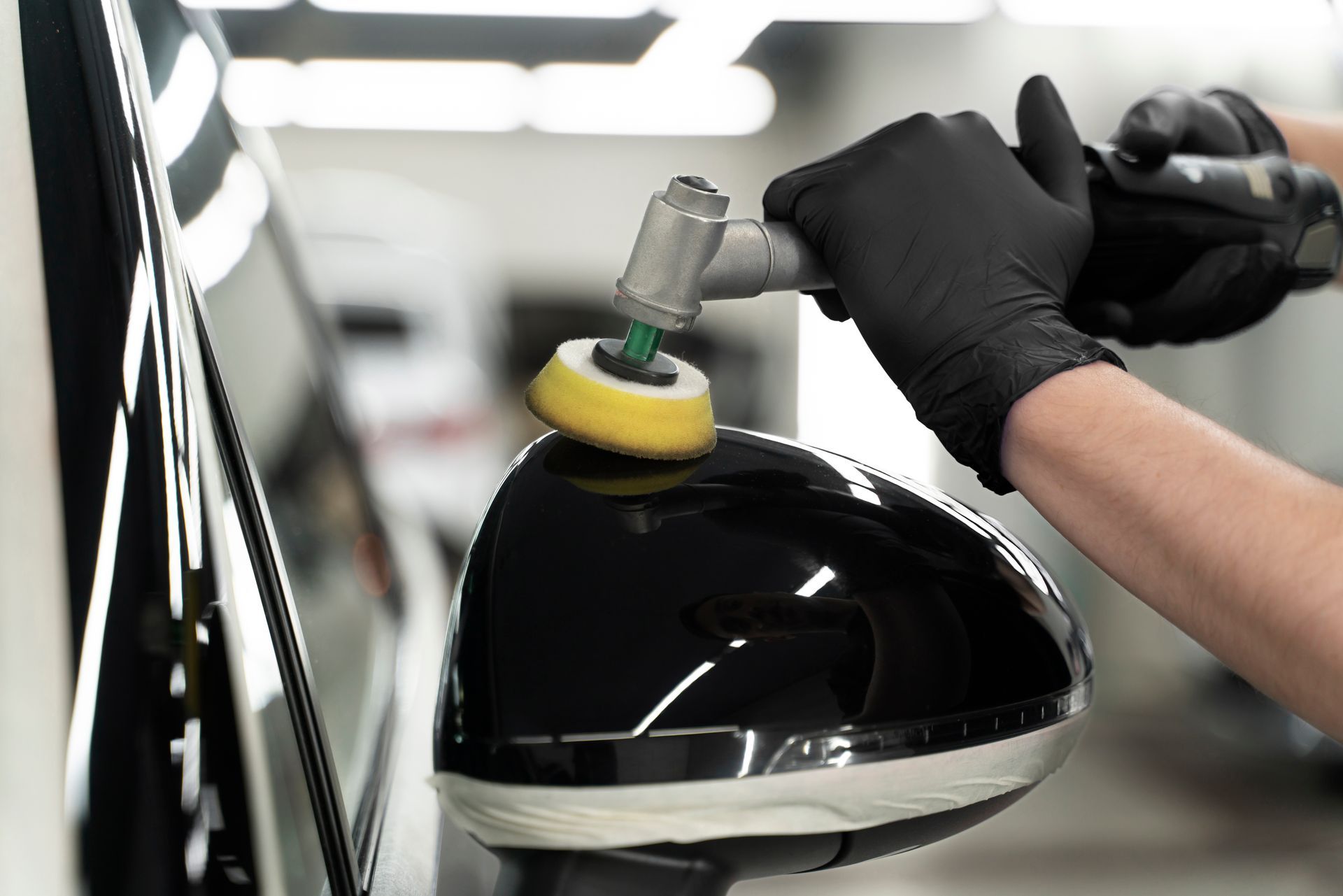 Gloved hand polishing a glossy black car side mirror with a buffer in a garage