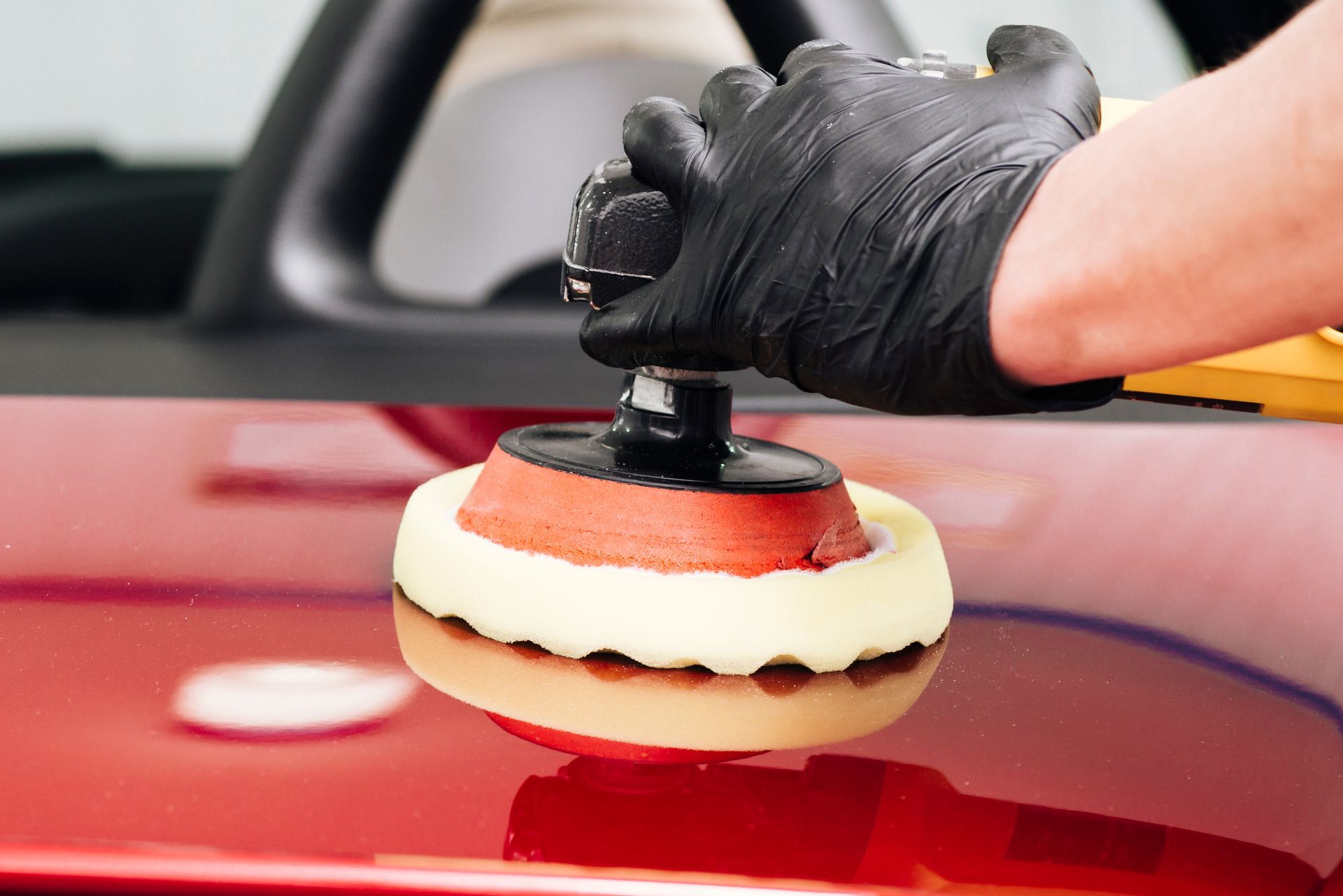 Gloved hand polishing a red car hood with a buffer pad