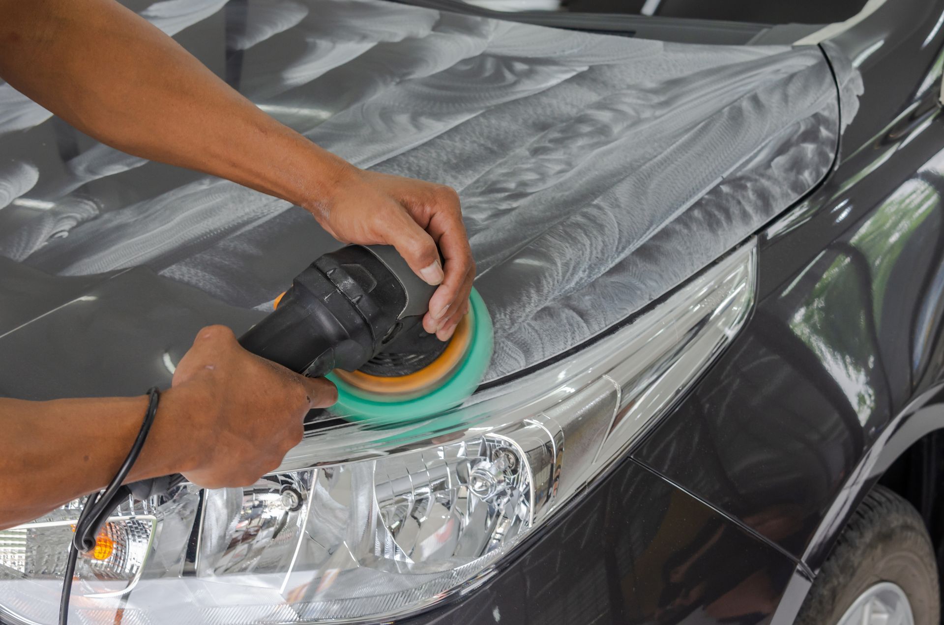 Hands polishing a black car headlight with a rotary buffer and green pad