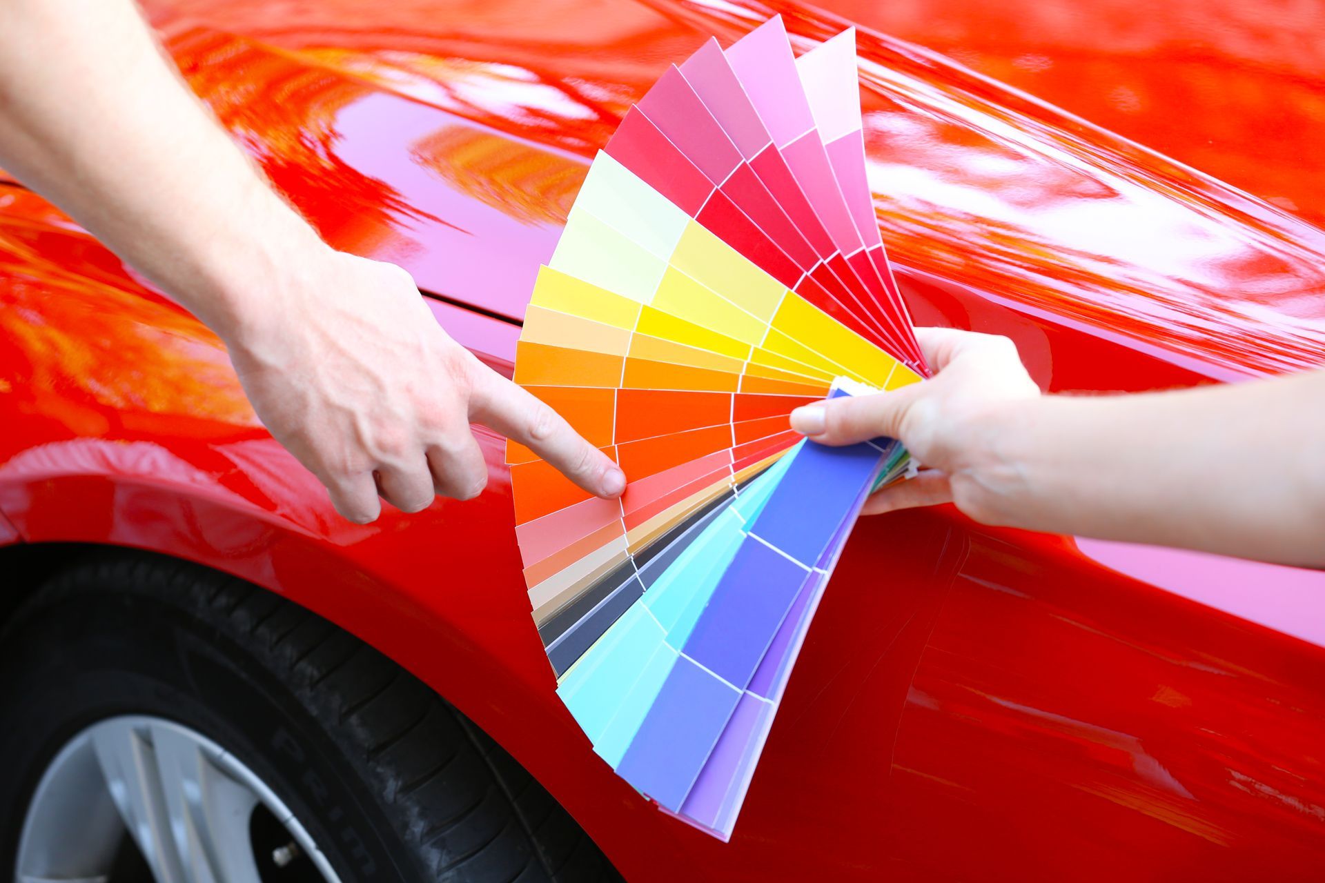 Hands pointing at a fan of paint color swatches on a red car door