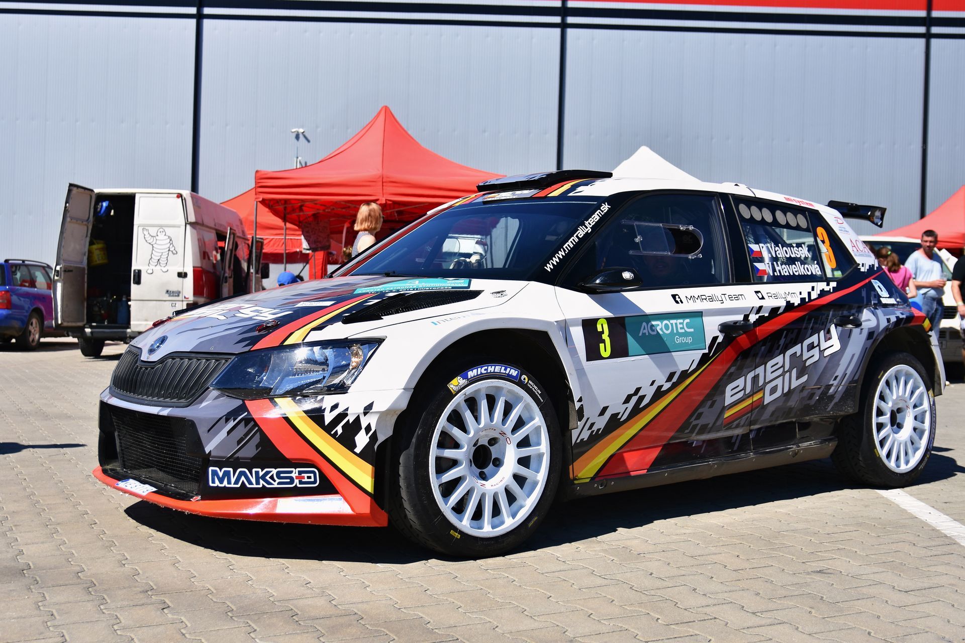 Rally car with white, black, and orange livery parked by a red tent at an event venue
