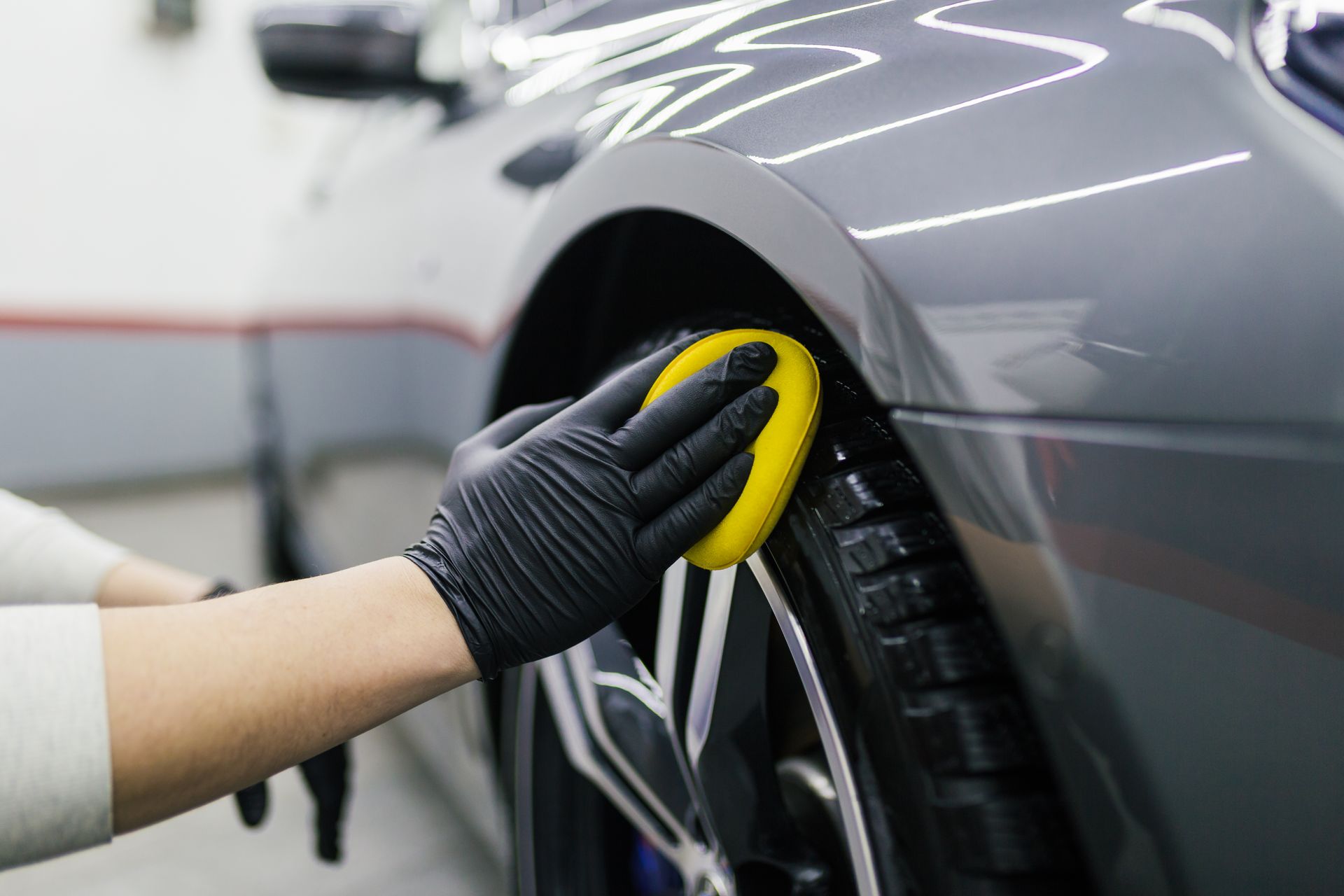 Gloved hand cleaning a car wheel with a yellow sponge in a garage