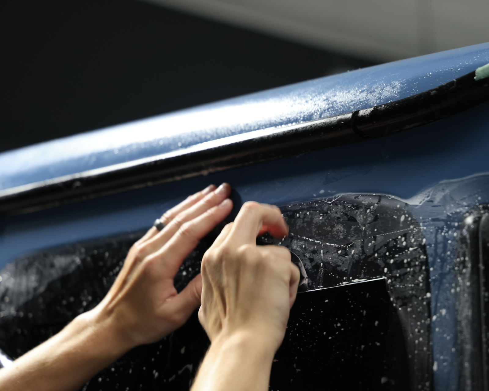 Hands gripping a wet car roof rack beside a tinted window, with sunlight reflecting off the black vehicle.