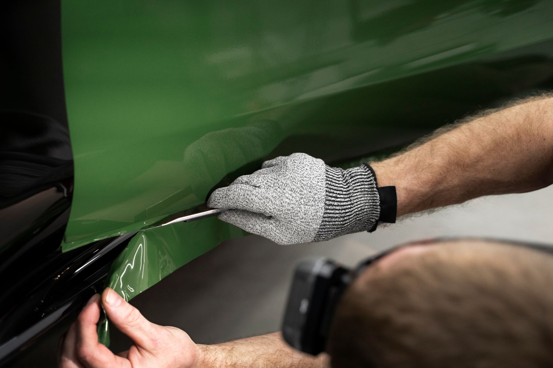 Gloved hands applying green vinyl film to a car panel with a squeegee tool