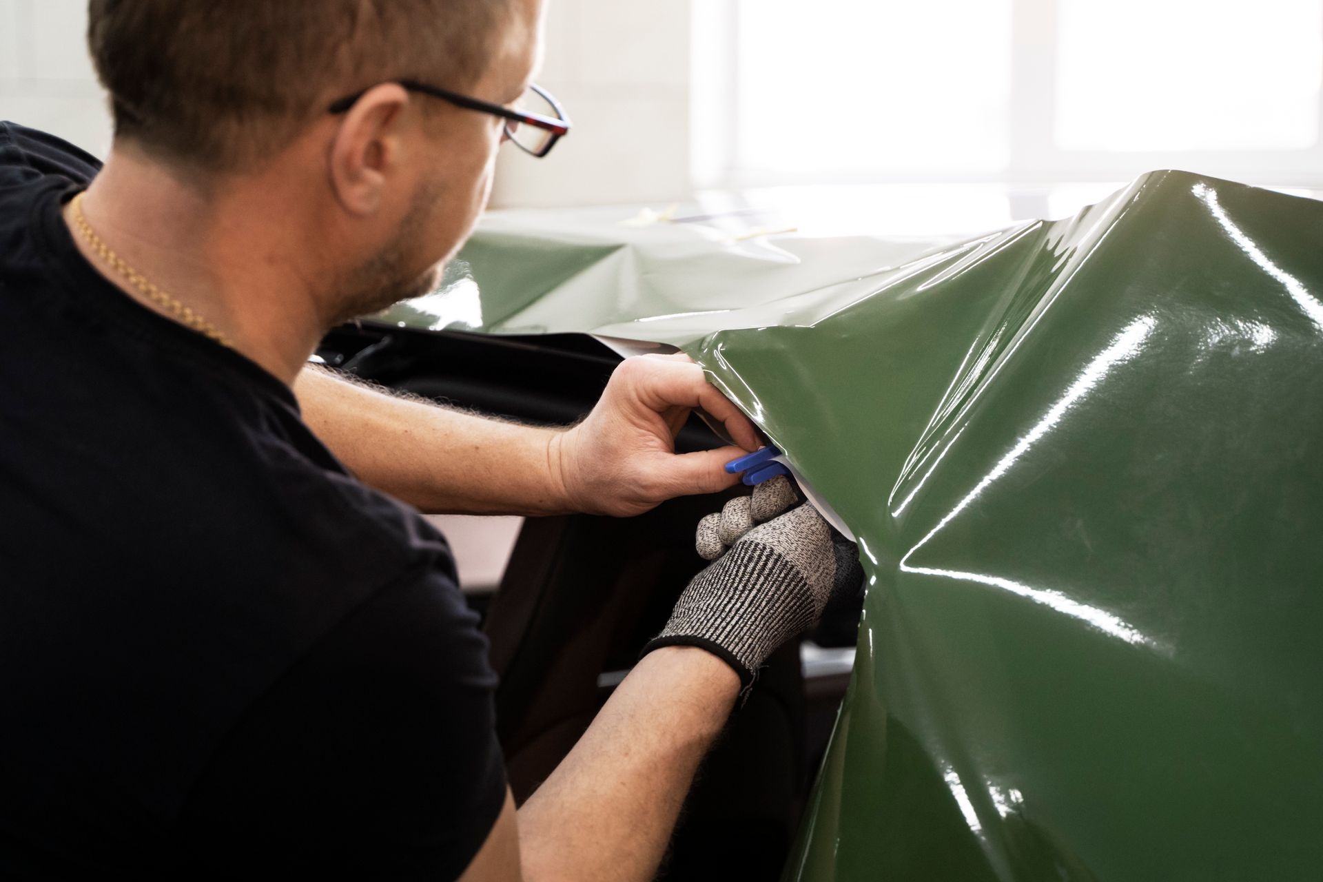 Person in gloves applying green vinyl wrap to a car panel with a tool