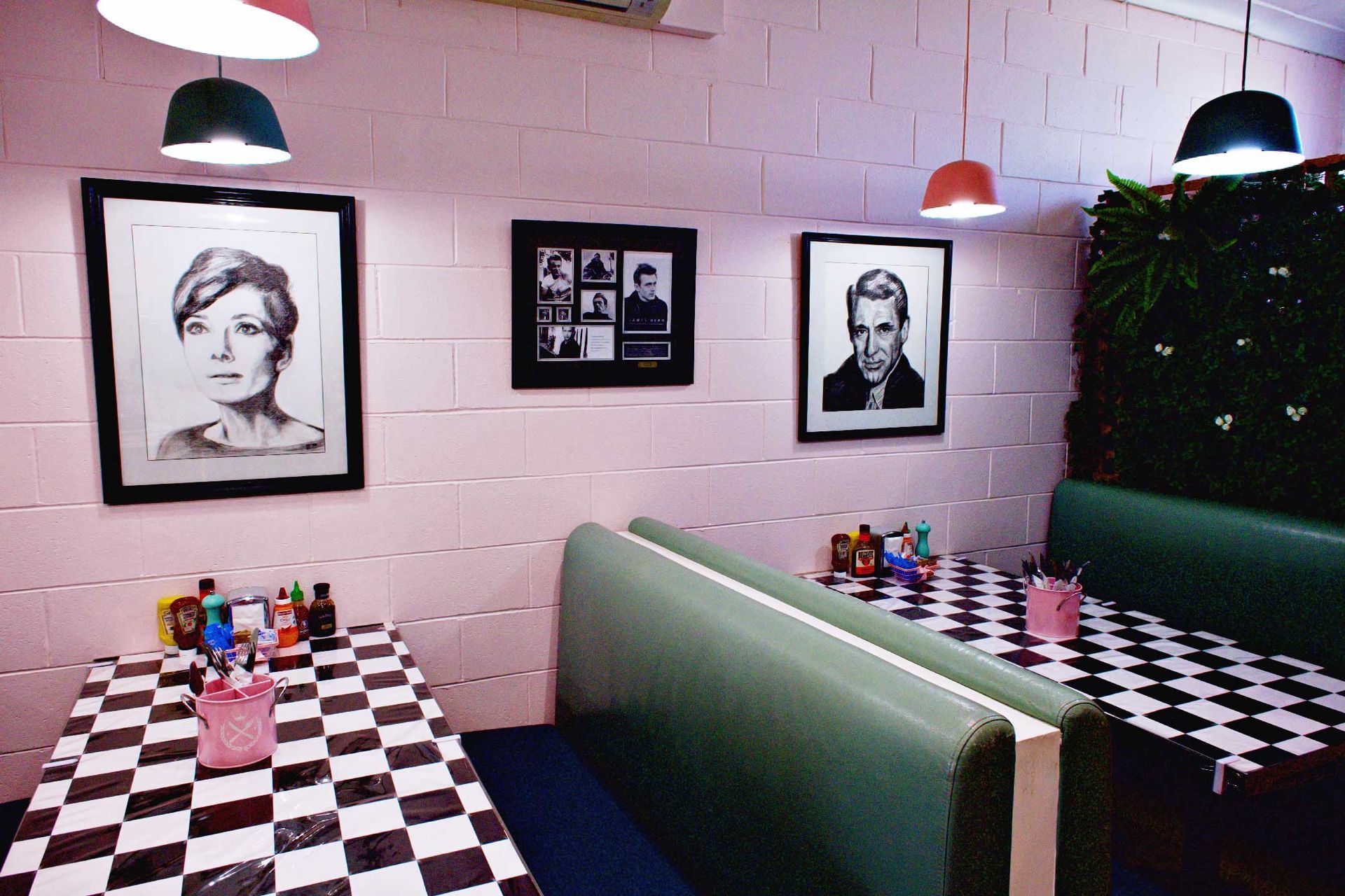 Diner interior with pink walls, black and white checkered tables, green booths, framed portraits, and hanging lights.