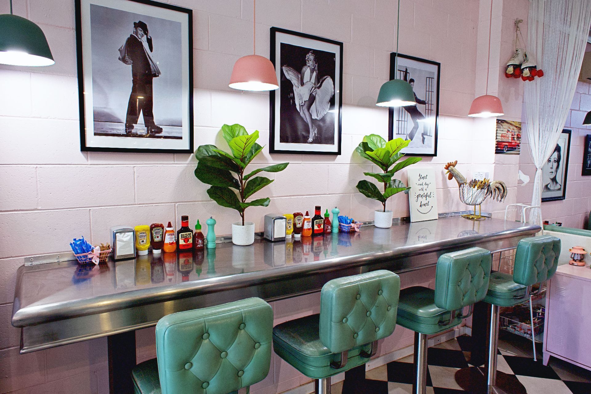 Diner counter with green stools, pink walls, framed art, potted plants, and various condiments.
