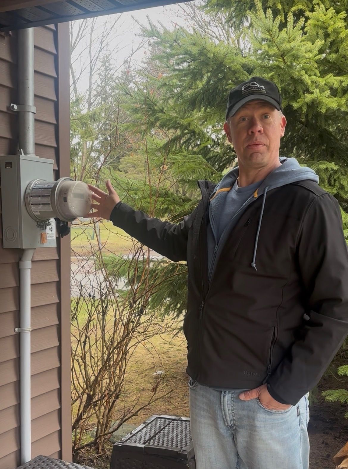 Man standing by a house meter box outdoors, hand on the electrical conduit, wearing a black jacket and cap.
