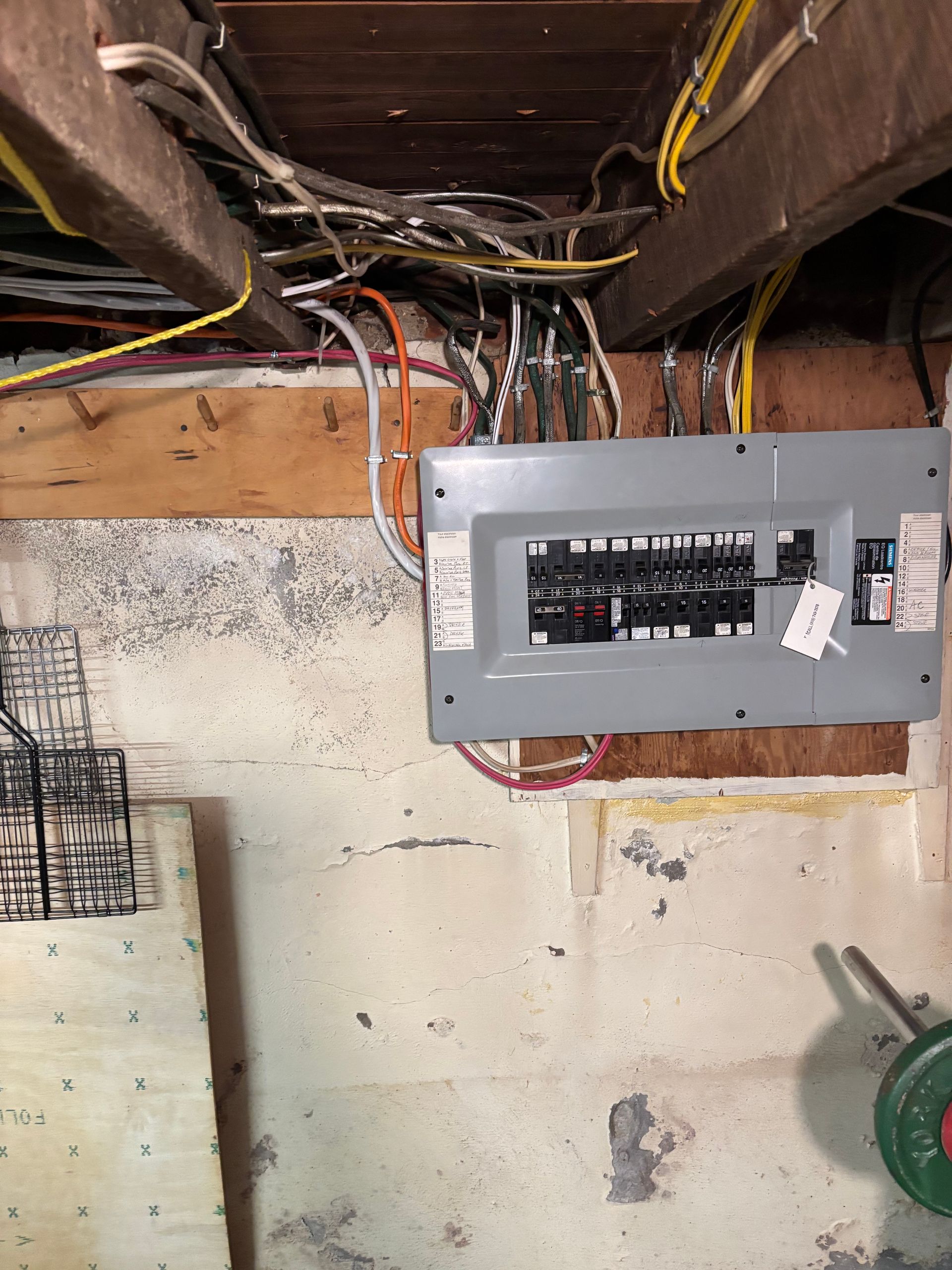 Electrical breaker panel mounted under exposed ceiling joists with visible wiring and unfinished wall.