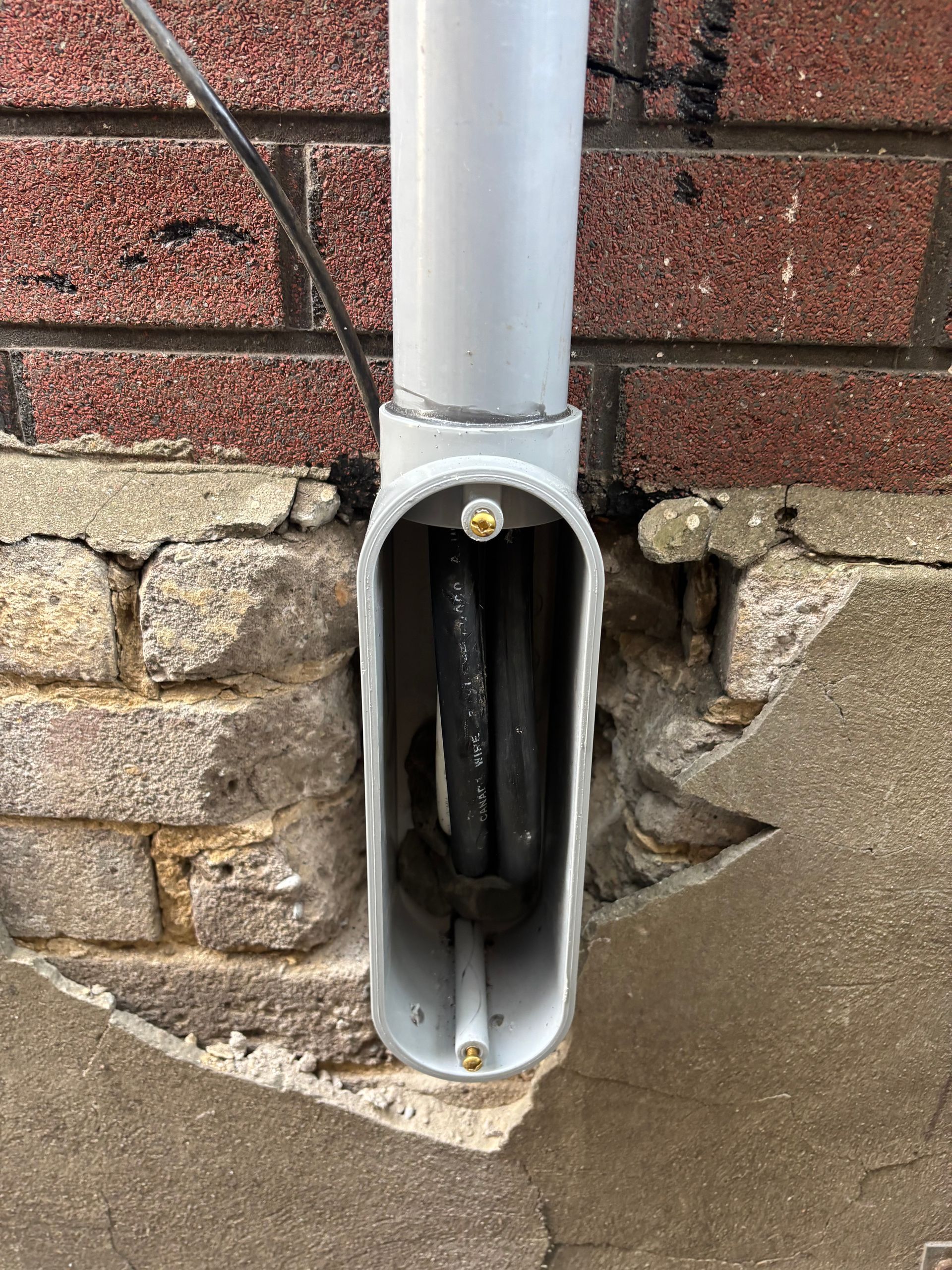 Open electrical outlet box in a brick wall with exposed wiring beside a downspout