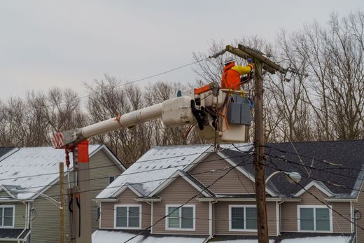 Utility workers repairing power lines from a bucket lift above snowy rooftops.