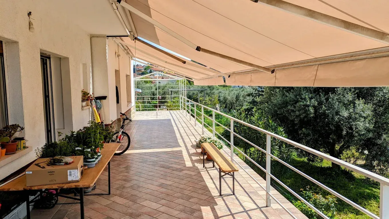 Passaggio pedonale esterno coperto con panchine, ringhiera, piante in vaso e vegetazione accanto a un edificio.