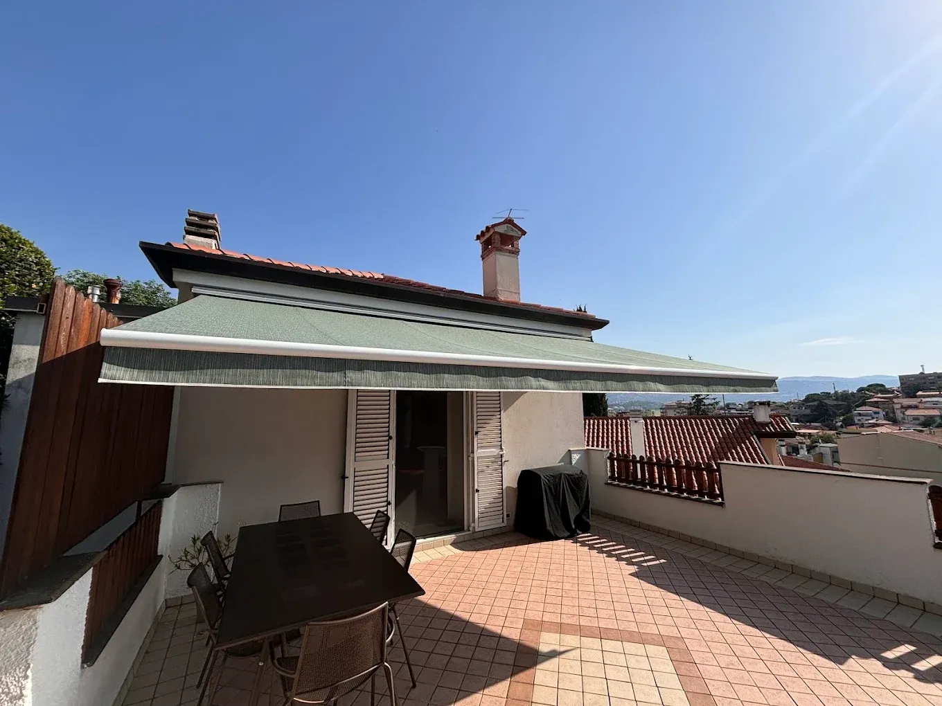 Terrazza panoramica soleggiata con tenda da sole, tavolo e sedie da giardino e vista sulla città sotto un cielo azzurro.