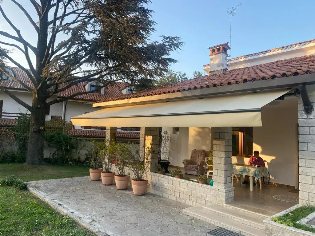 Patio sul retro con tenda da sole, piante in vaso e una persona seduta vicino alla casa al crepuscolo.