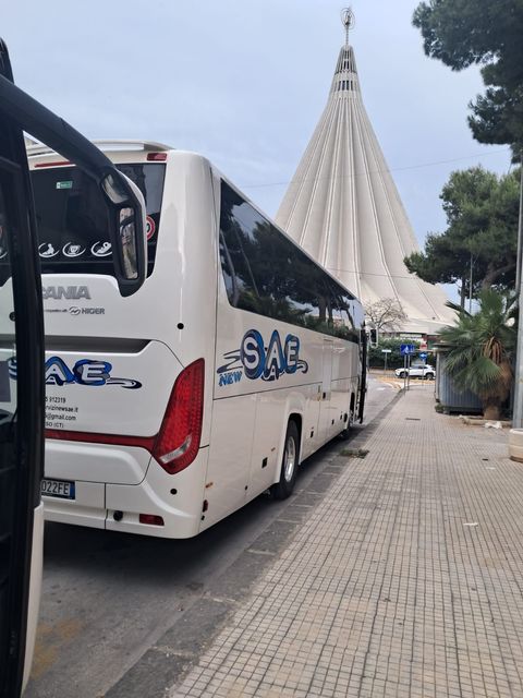 Autobus turistico bianco parcheggiato su una strada acciottolata accanto a un grande grattacielo moderno, sotto un cielo nuvoloso