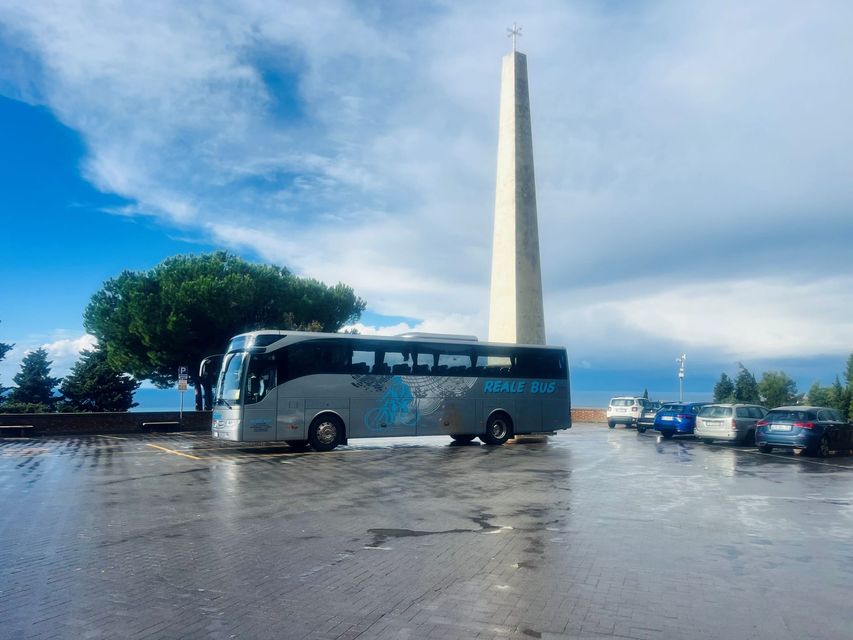Pullman turistico vicino a un alto obelisco su una piazza bagnata, sotto un cielo azzurro nuvoloso