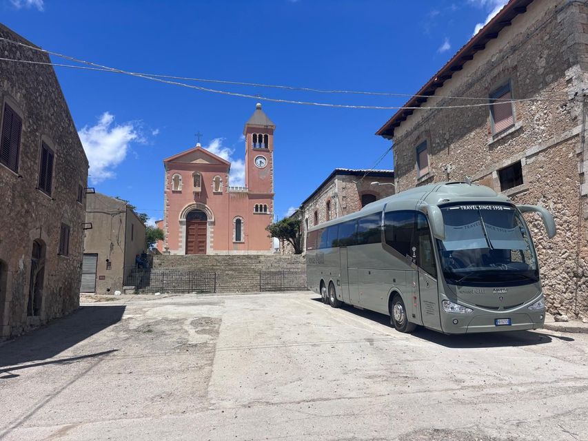 Cortile in pietra con una chiesa in mattoni e un campanile sotto un cielo azzurro, con un autobus argentato parcheggiato sulla destra.