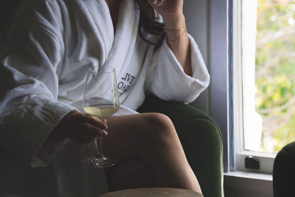 Person in white robe sitting by a window, holding a wine glass and reading a magazine.