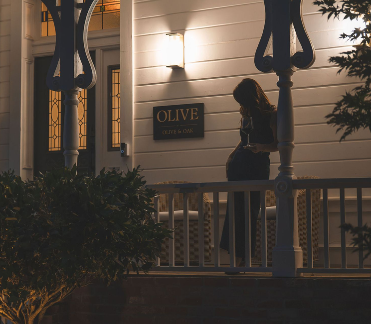 Woman standing on a porch by a lit wall sign reading &ldquo;Olive&rdquo; at dusk