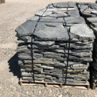 Stack of gray flagstone on a wooden pallet, secured with black straps, outdoors on a sunny day.
