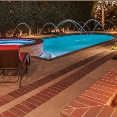 Swimming pool with fountains at night; brick patio and hot tub visible.