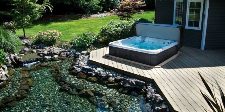 Hot tub on a wooden deck next to a small stream. Lush green yard surrounds the deck and house.