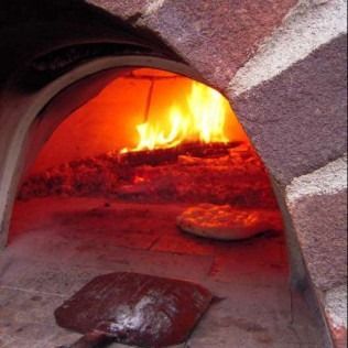 Wood-fired oven interior, flames burning, pizza cooking. A pizza peel rests in foreground.