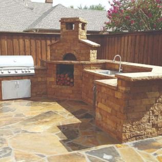 Outdoor kitchen with a stone pizza oven, grill, sink, and wood storage, on a stone patio.