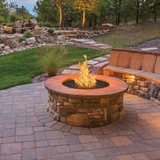 Fire pit on a stone patio with a built-in stone bench, steps, and landscaping in the background.