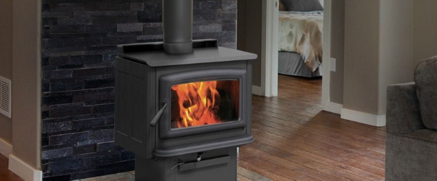 A burning wood stove in a living room, with a dark stone accent wall, wooden floors, and a glimpse of a bedroom.