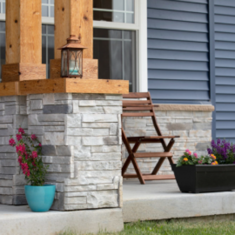 Stone-covered porch with wood columns, blue siding, a chair, and flower pots.