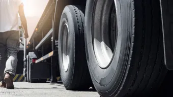 Person walking beside a truck with large black tires on a sunny street