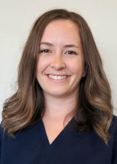 Headshot of a person with shoulder-length wavy brown hair, smiling warmly, wearing a dark blue v-neck scrub top.