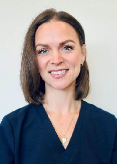 A person with shoulder-length brown hair wearing a dark blue v-neck scrub top and a small pendant necklace.